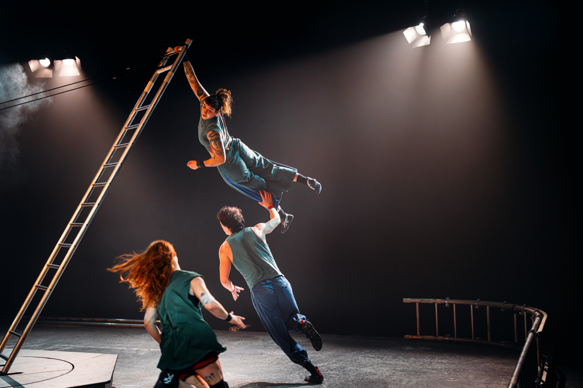Aurora Mesropyan in BEYOND: she hangs from the top of a tall ladder, held up from below by a base, while a third performer runs in
