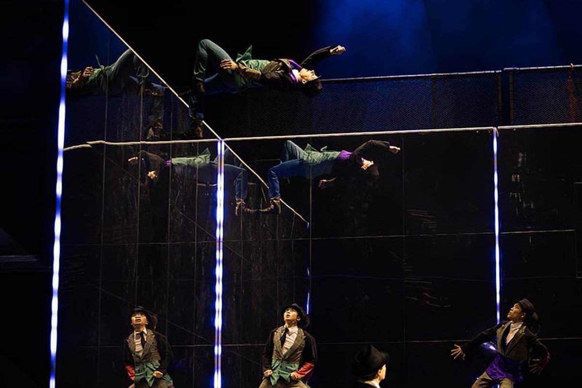 Two ALIZÉ acrobats flying horizontally along a trampwall, mirrored in a diagonal glass panel, with performers in bowler hats looking up from the stage below.
