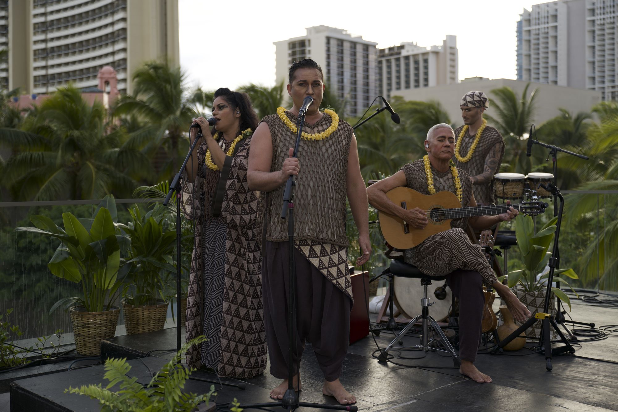 ʻAuana's singers and musicians performing on an outdoor stage in Waikīkī: two singers at microphones in patterned costumes and yellow lei, a guitarist and a percussionist behind them, with palm trees and hotel towers in the background.