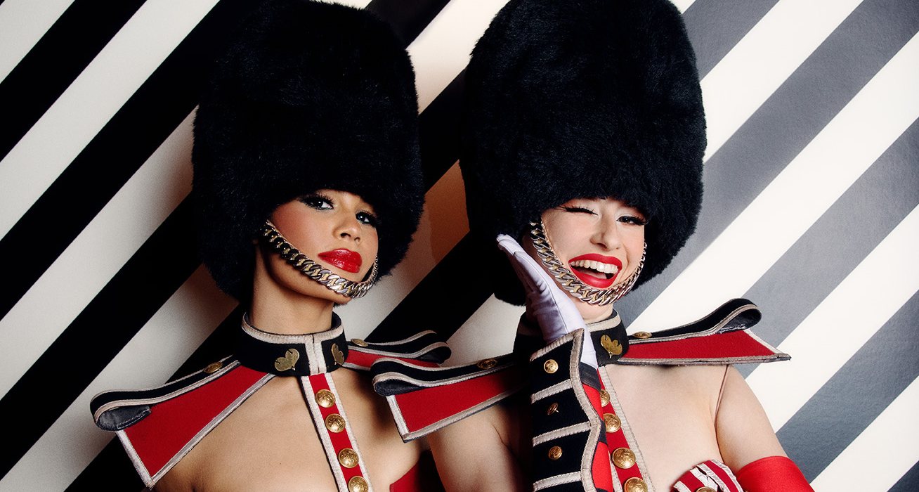 Two Crazy Horse dancers in guardsman costumes and bearskin hats, one winking, against a black and white striped wall