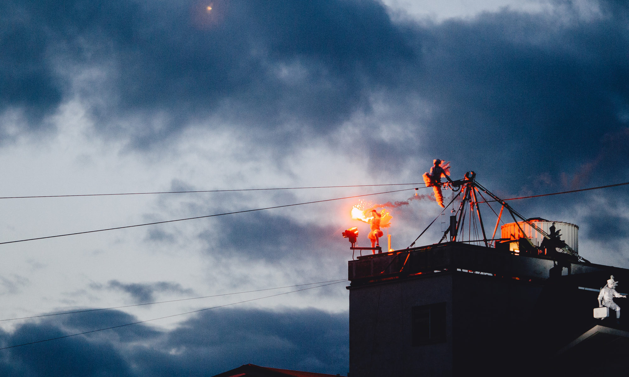 Dusk over a rooftop: one performer in a feathered costume holds a burning flare, another hangs from a metal rig above the street wires.