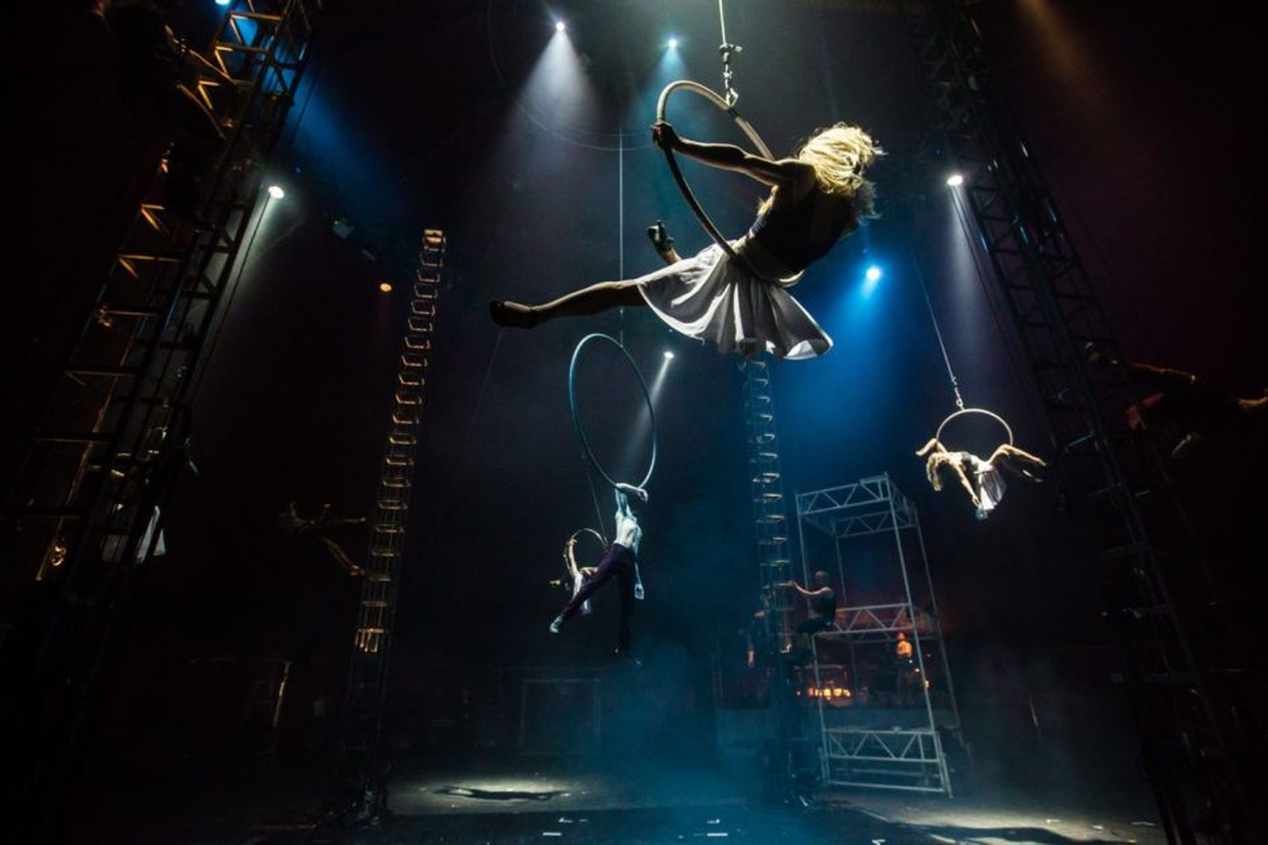 NoFit State Circus aerialists on hoops at different heights under blue stage lights, one in a white skirt swinging in the foreground.