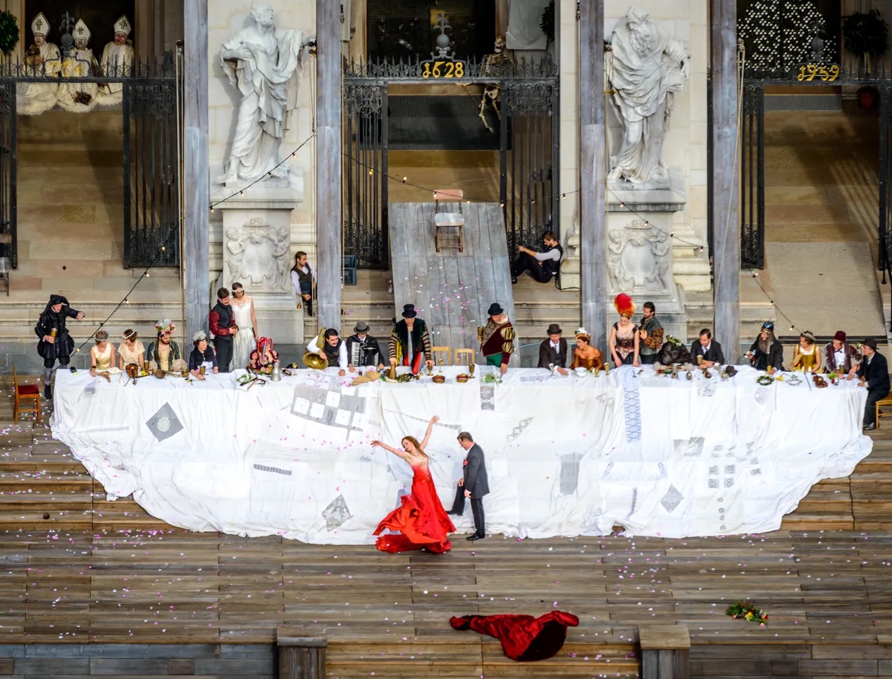An open-air performance on a wooden stage in front of a cathedral facade: a woman in a red dress dances below a long banquet table lined with costumed actors.