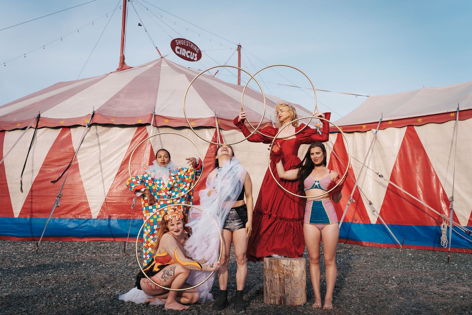Five Shoestring Circus performers in costume holding gold hoops in front of the company's red and white striped big top, under a blue sky.