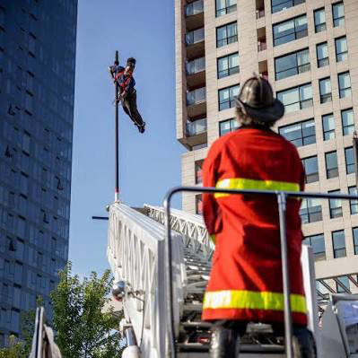 A performer hangs from the extended ladder of a fire truck above a crowd in Montreal, the observation wheel behind