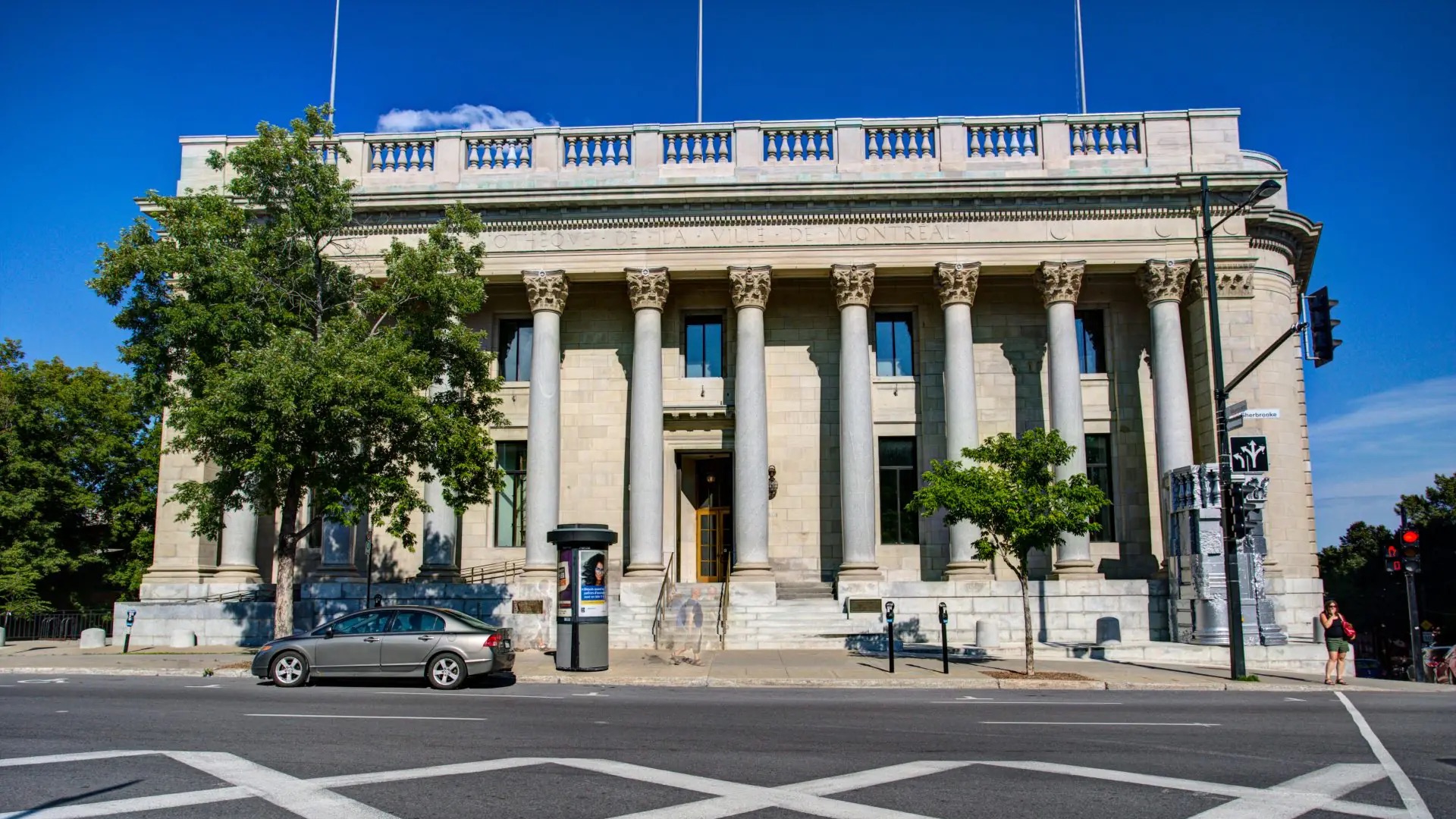 The neoclassical stone facade of the Bibliothèque de la Ville de Montréal on Sherbrooke Street, columns and balustrade against a clear blue sky.