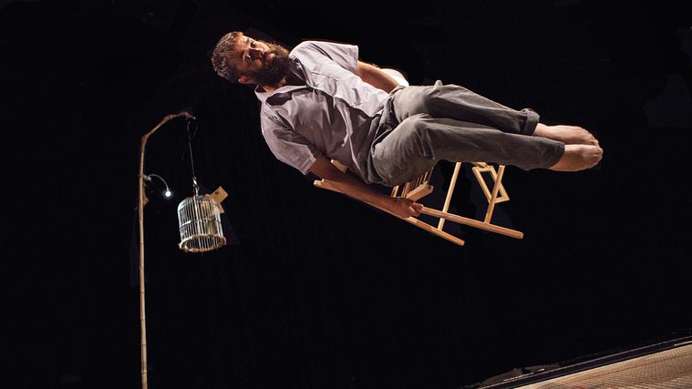 A performer balances horizontally across a folding wooden chair on a dark stage, a birdcage hanging from a bamboo lamp nearby
