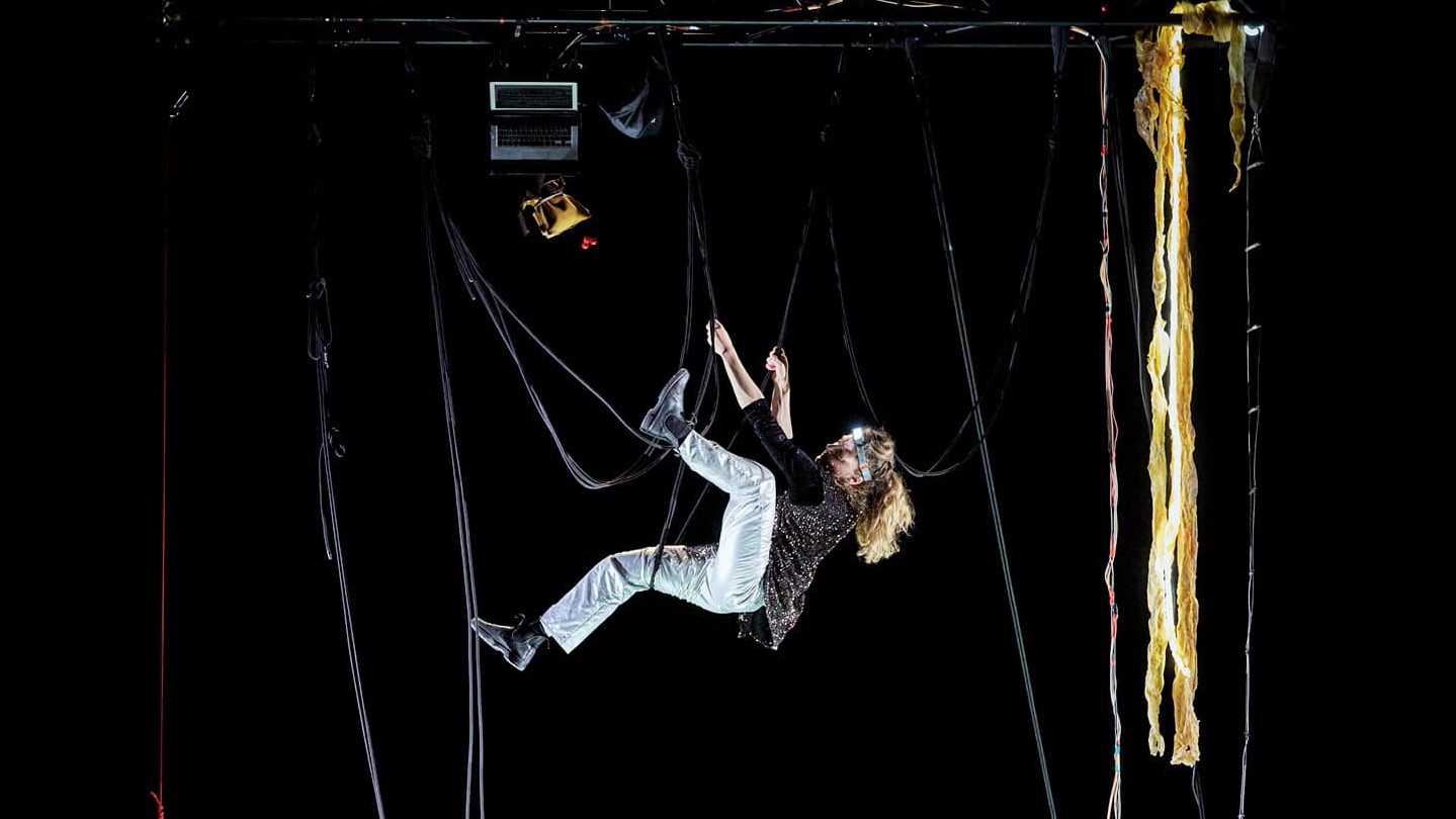A performer hangs in a web of black ropes in a darkened space, wearing a head torch.