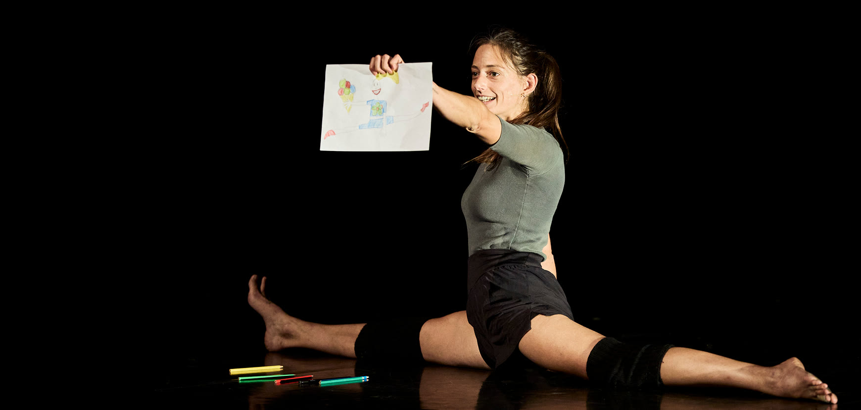A circus artist in the splits on a dark stage, smiling and holding up a child's drawing