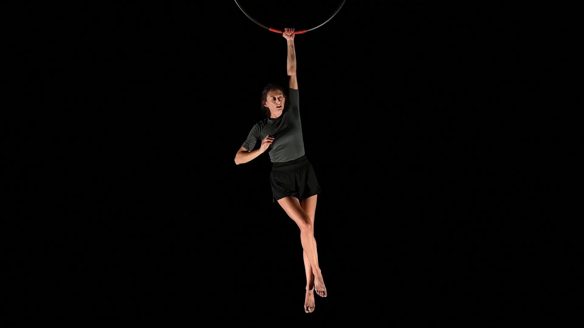 Aurora Dini hanging by one hand from an aerial hoop against a black background