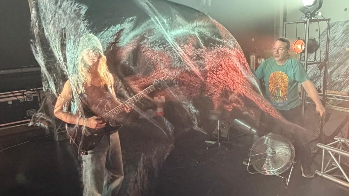 A guitarist and a seated musician under a billowing sheet of printed fabric on a lit rehearsal stage.