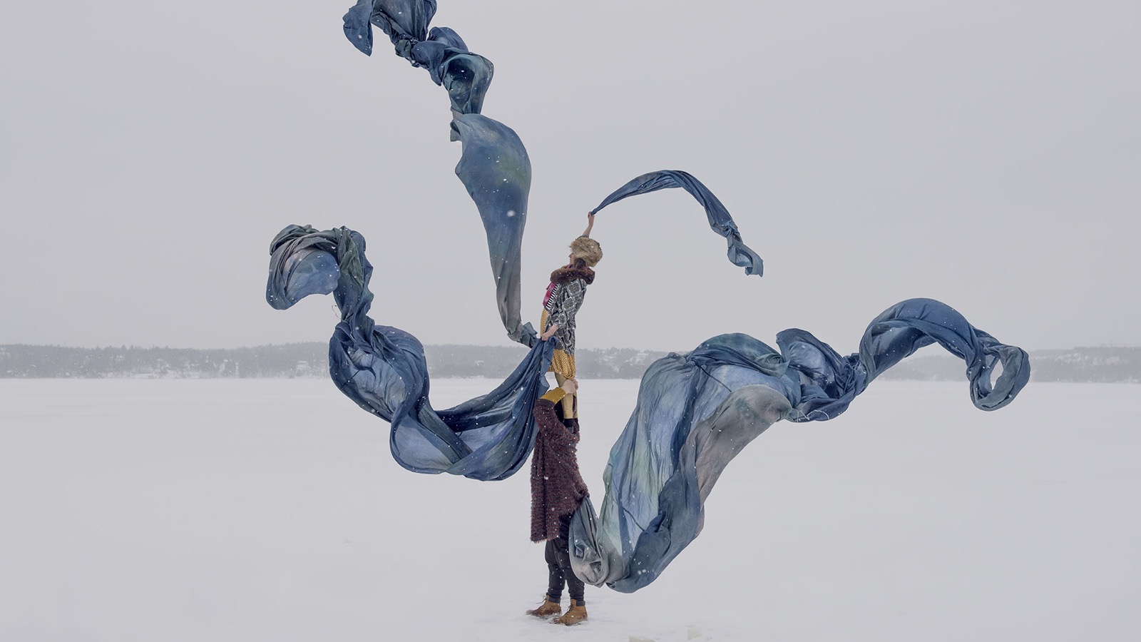 One performer stands on another's shoulders on a frozen lake, long blue silks swirling around them