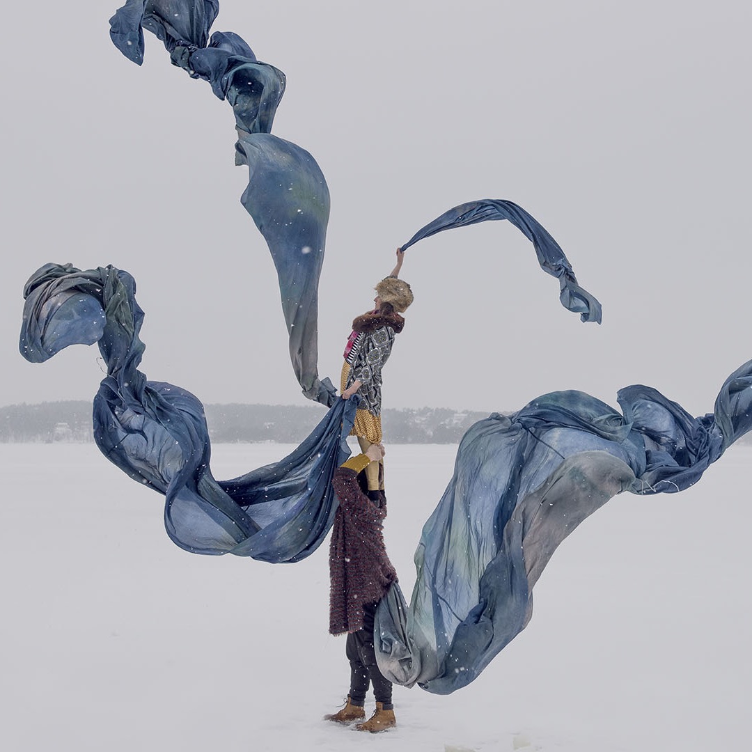 One performer stands on another's shoulders on a frozen lake, long blue silks swirling around them