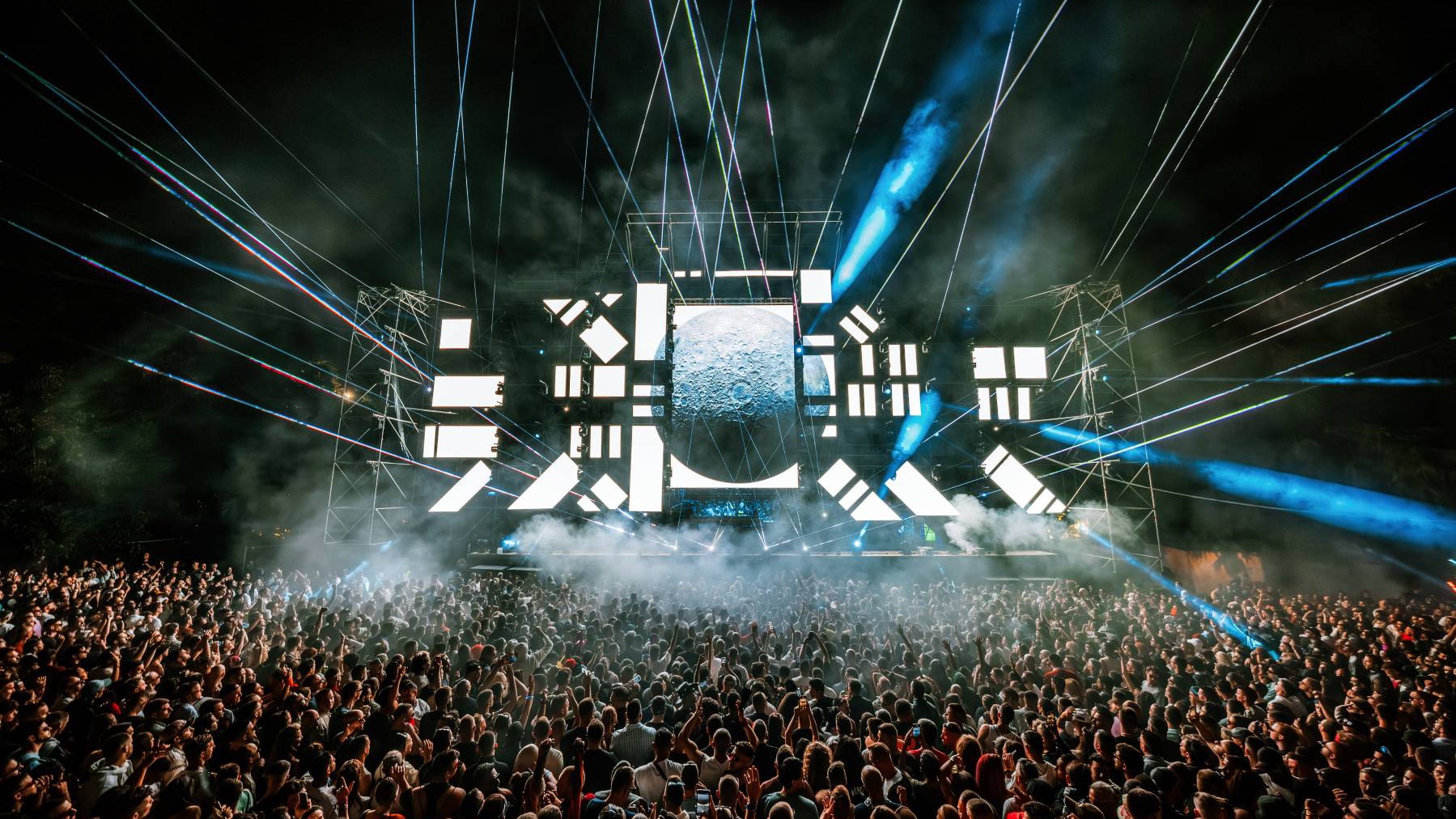A night-time festival crowd in front of a stage lit by lasers and white LED panels