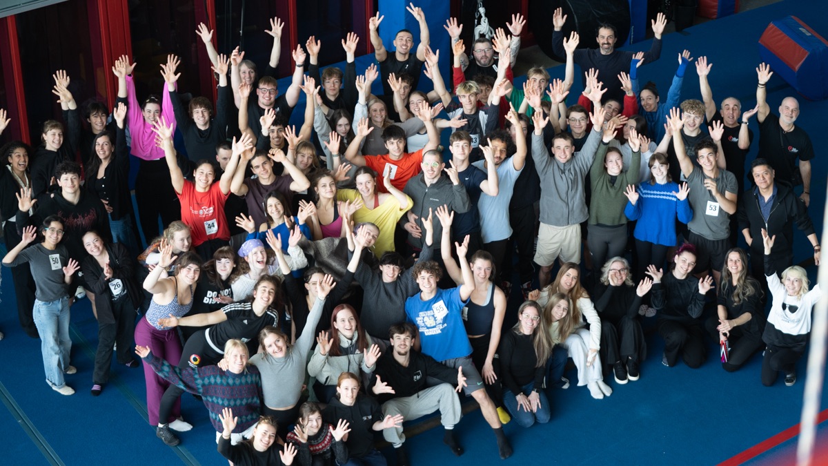 Around a hundred candidates stand together on blue gym mats with their arms raised, audition numbers pinned to their clothes.