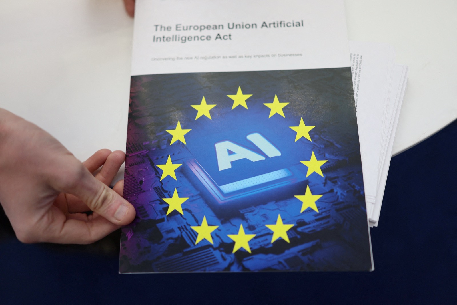 A hand holds a printed copy of the European Union Artificial Intelligence Act, its cover showing the EU stars circling a chip