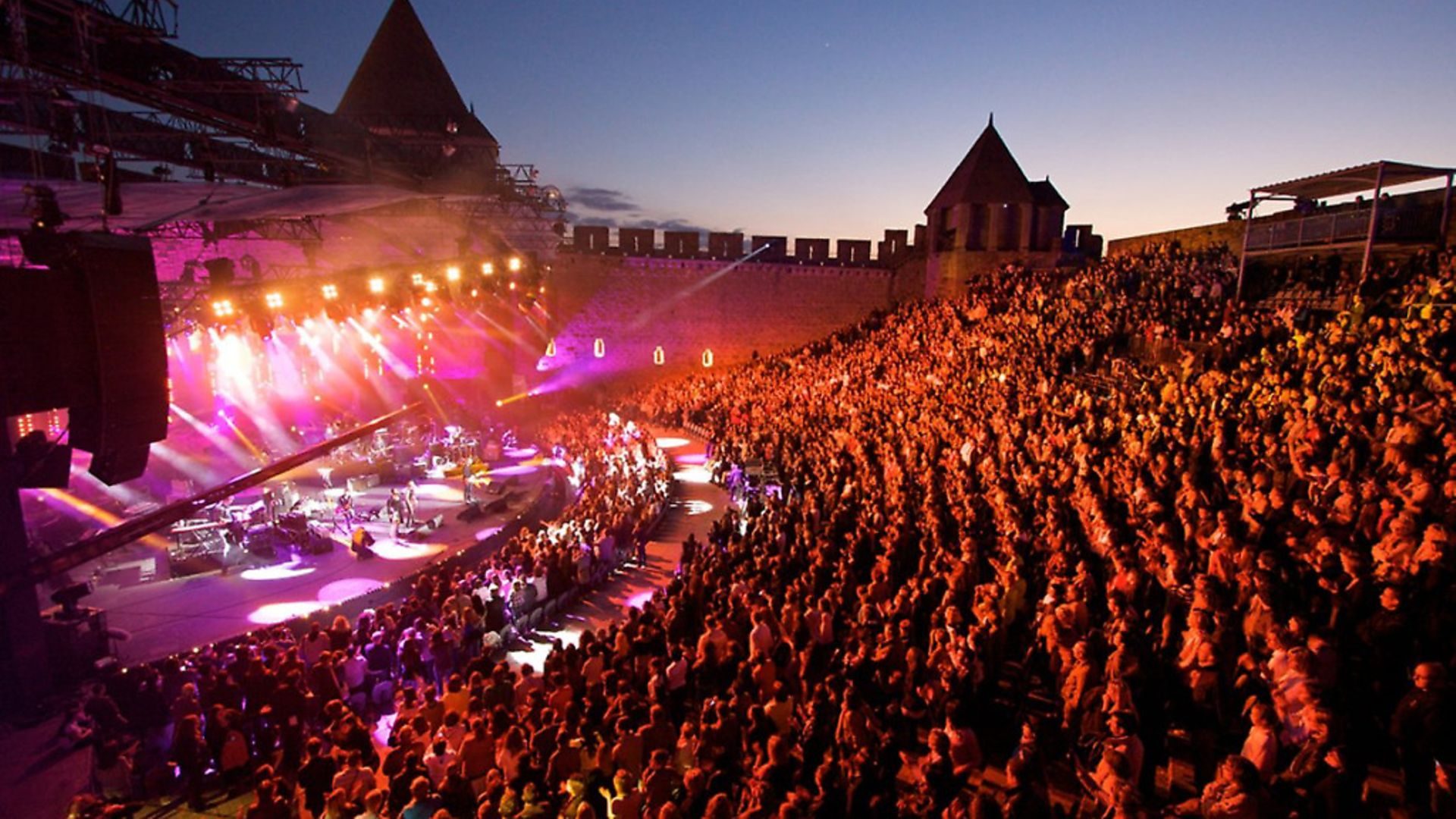 A large crowd fills a fortress amphitheatre at dusk in front of a lit concert stage