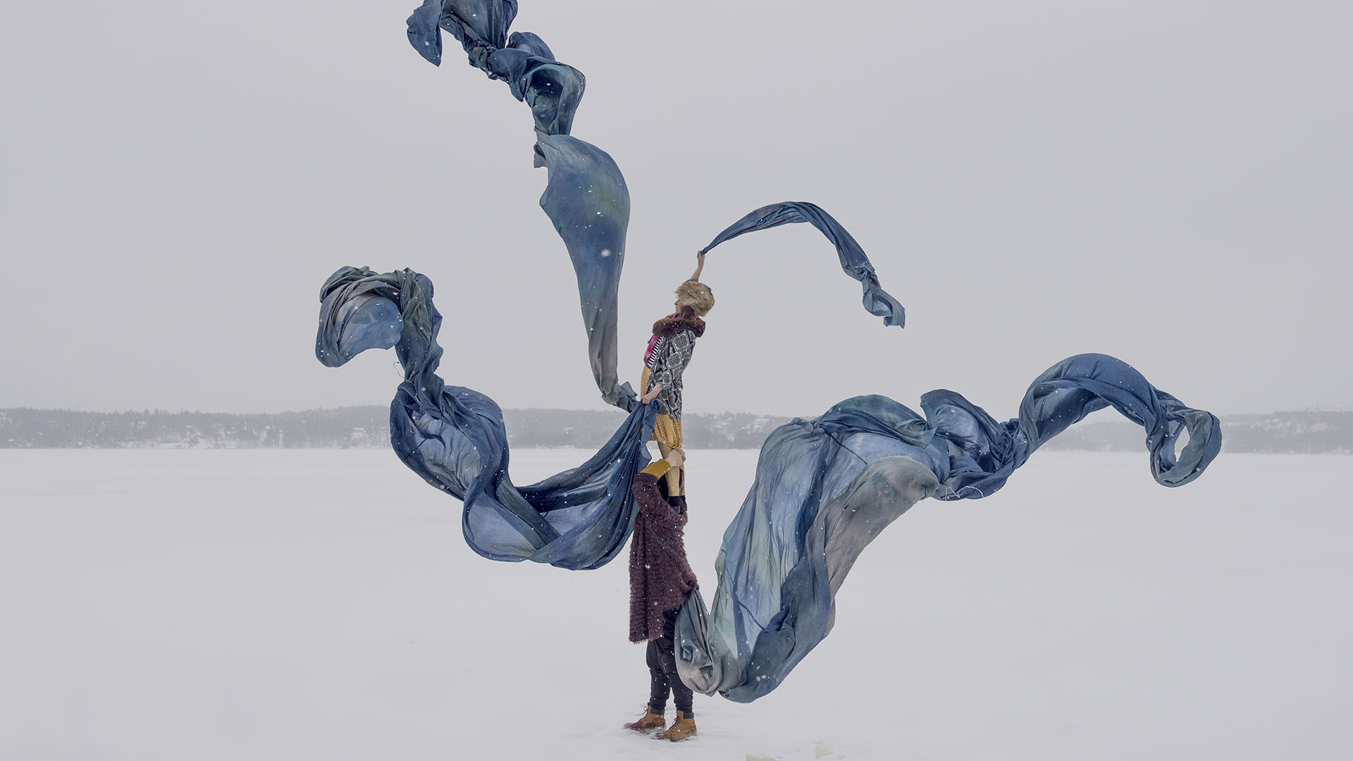 One performer stands on another's shoulders on a frozen lake, long blue silks swirling around them