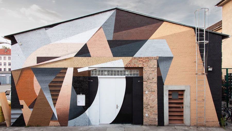 The Katapult building in Berlin Oberschöneweide, its brick facade painted with a large geometric mural in pale blue, peach and black, with white double doors at the centre