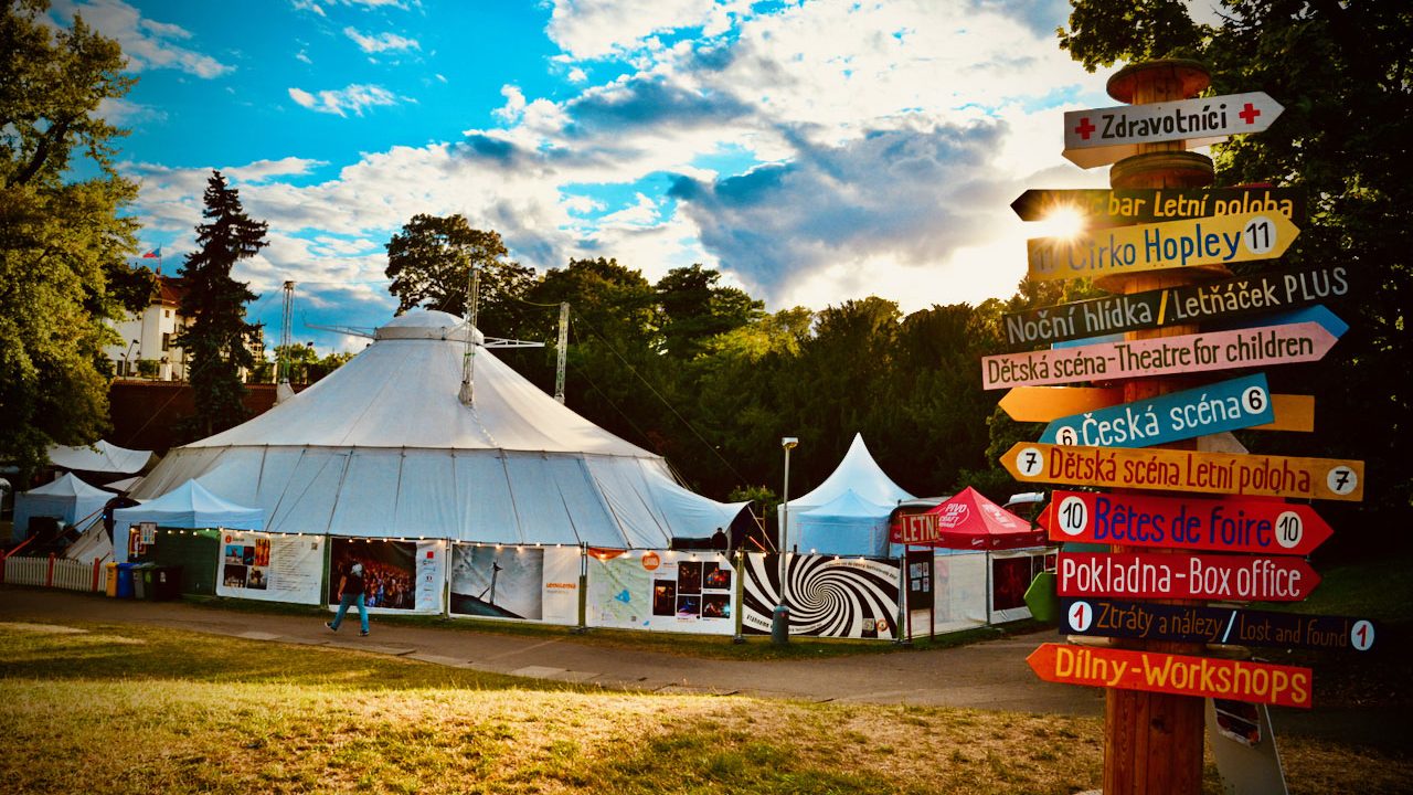 The white Letní Letná big top in a Prague park at golden hour, beside a signpost pointing to the box office, workshops and children's stage