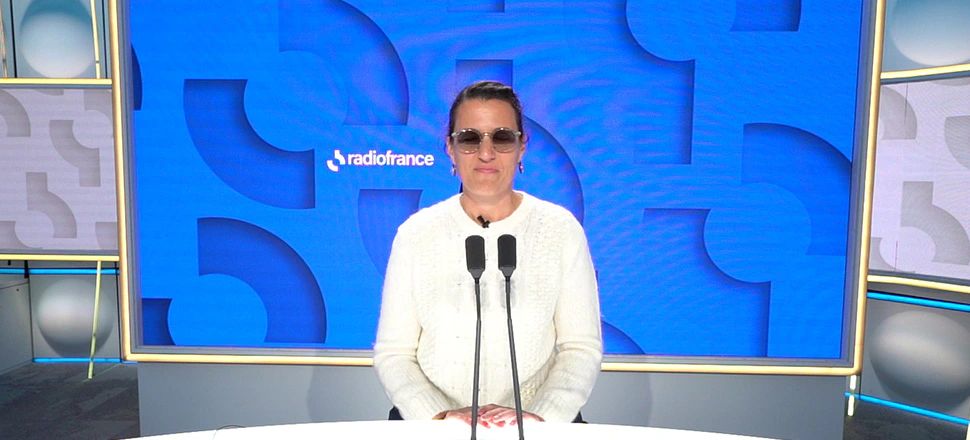 Lætitia Bernard seated at two microphones in a Radio France studio, wearing tinted glasses and a white jumper