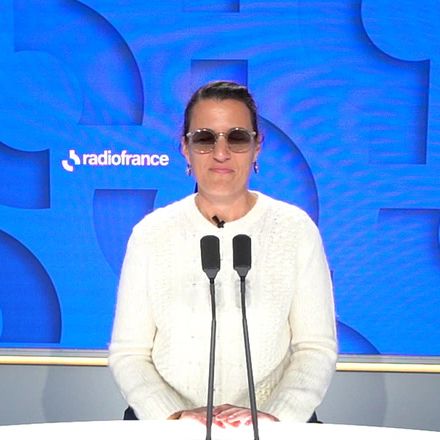 Lætitia Bernard seated at two microphones in a Radio France studio, wearing tinted glasses and a white jumper