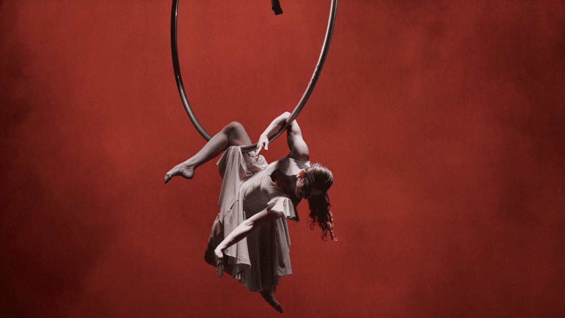 An aerialist in a white dress hangs upside down from an aerial hoop against a deep red background.