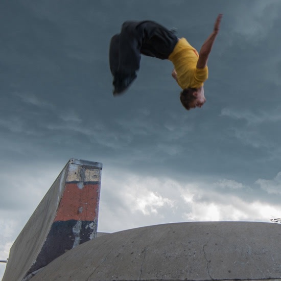 A rider backflips above a skate park ramp while teenagers with scooters watch under a grey sky.