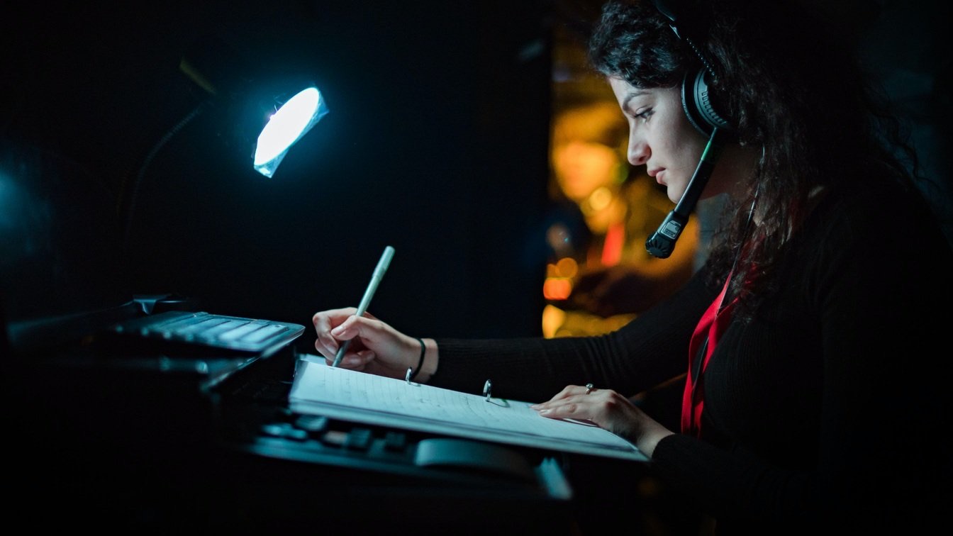 Someone on a headset writing in a prompt book under a desk lamp in a dark booth