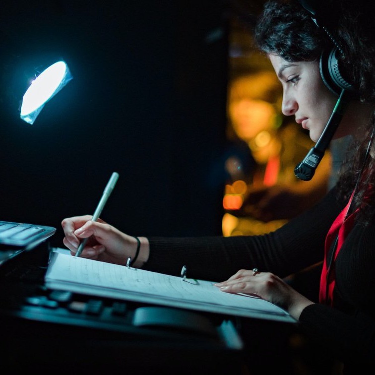 Someone on a headset writing in a prompt book under a desk lamp in a dark booth