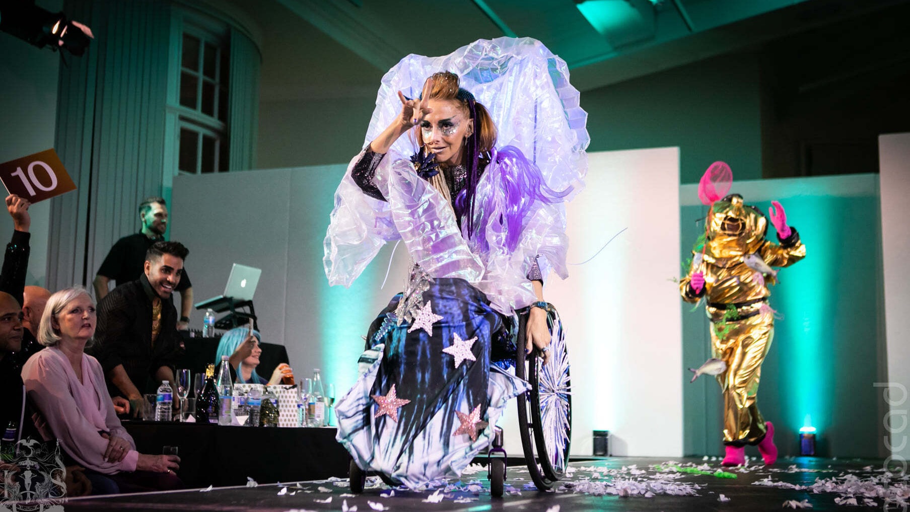A performer in an iridescent inflated costume rides a decorated wheelchair down a catwalk, a judge holding up a score of 10 at the side.